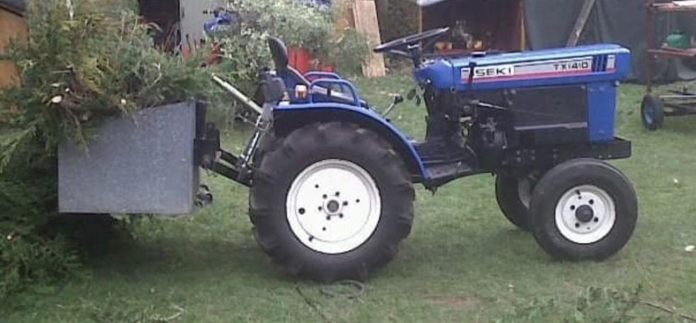 Blue tractor with a metal attachment, parked on grass near a shed.